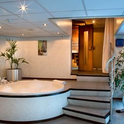 Modern wellness area on SE Manon ship with round white whirlpool, wooden sauna, exercise bike, and decorative plants under ceiling lighting.