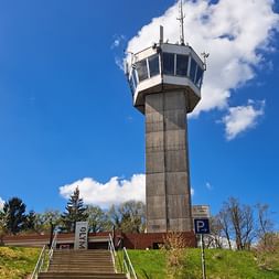 Betonaussichtsturm Deutscher Olymp mit gläserner Aussichtskabine an der Spitze, Treppe führt einen grasbewachsenen Hügel hinauf, blauer Himmel mit weißen Wolken.