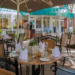 Außenterrasse im Upstalsboom Parkhotel Emden mit gedeckten Tischen, Korbstühlen unter Sonnenschirmen, umgeben von Grün und Hotelgebäude.