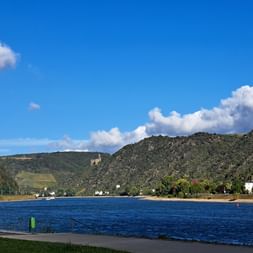 Breiter Fluss zwischen grünen Hügeln mit Weinbergen bei St. Goarshausen. Weiße Häuser am rechten Ufer unter blauem Himmel.