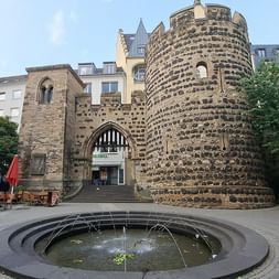 Mittelalterlicher Steinturm mit rundem Turm im Bonner Stadtzentrum. Runder Brunnen im Vordergrund, rote Sonnenschirme und Außenbereich sichtbar.