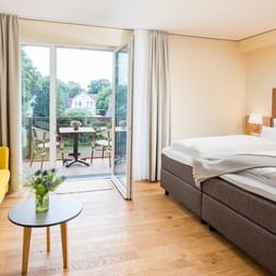 Helles Hotelzimmer im Parkhotel Jever mit Doppelbett, gelbem Sofa, Holzboden und Balkontür zu einem Sitzbereich im Freien.