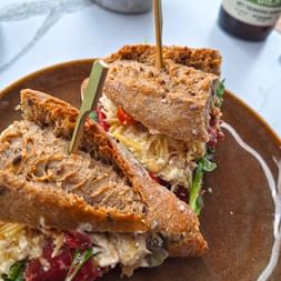 Gestapeltes Sandwich mit Mehrkornbrot, Käse, Gemüse und Fleisch auf einem braunen Teller. Sirup wird über das Sandwich gegossen.