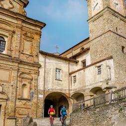 Zwei Radfahrer in bunten Trikots fahren durch den historischen Burghof von Cravanzana mit Steinkirchenfront und mittelalterlichem Turm.