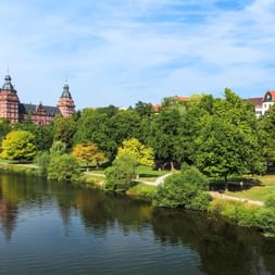 Blick auf Schloss Johannisburg in Aschaffenburg vom Main aus, zeigt rotes Sandsteinschloss mit Türmen umgeben von grünen Bäumen.
