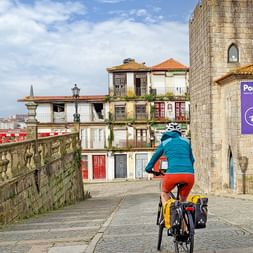 Radfahrer mit blauer Jacke und roter Hose fährt auf Kopfsteinpflaster in Portos historischem Viertel mit bunten Häusern und Steinturm.