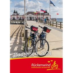 Zwei Fahrräder mit roten Körben auf einer Holzseebrücke vor einem weißen historischen Gebäude mit Flaggen. Blauer Himmel über Sandstrand.