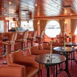 Eleganter Bar-Salon auf der MS Switzerland mit Holzvertäfelung, runden Bullaugen mit Vorhängen, gepolsterten Sitzgelegenheiten und Tresen.