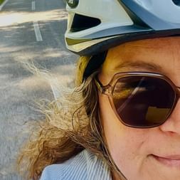 Nahaufnahme-Selfie einer lächelnden Radfahrerin mit Sonnenbrille und weißem Helm auf einem asphaltierten Radweg mit grünem Gras.