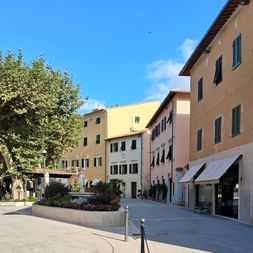 Fußgängerplatz in Casciani Terme mit bunten italienischen Gebäuden in Gelb, Rosa und Beige. Grüne Fensterläden, Markisen und Baum mit Blumenbeet.