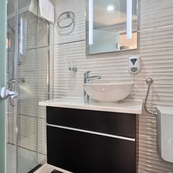 Modernes Badezimmer auf MY Magellan mit Glasdusche, weißem Waschbecken auf dunklem Unterschrank, strukturierten Wandfliesen und WC.