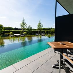 Moderne Terrasse mit Holztisch und Stühlen mit Blick auf einen Naturpool mit Wasserpflanzen im Hotel Zollhaus in Rastede.
