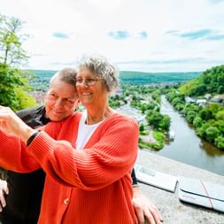 Glückliches Paar macht Selfie mit rotem Handy an malerischem Aussichtspunkt über Hann. Münden mit Weser und grünen Hügeln im Hintergrund.