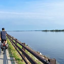 Zwei Radfahrer auf einer Brücke mit Holzgeländer am Alpe Adria Radweg bei Grado, mit ruhiger Lagune und ferner Küstenlinie unter blauem Himmel.