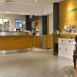 Moderne Hotelrezeption mit Holztresen und Wand mit Landhotel Schnuck Logo. Brauner Sessel und weißer Schrank mit Broschüren im Vordergrund.