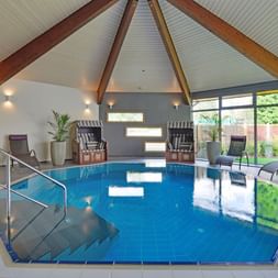Hallenbad im Landhotel Schnuck mit gewölbter Holzdecke, Liegestühlen, Strandkörben und großen Fenstern mit Blick auf den Garten.