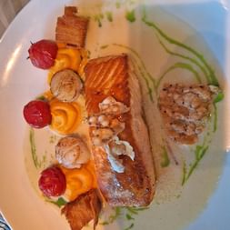Gegrilltes Lachsfilet mit gebratenen Jakobsmuscheln, Kirschtomaten, Pfifferlingen und knusprigem Schweinebauch auf weißem Teller mit Sahnesauce.