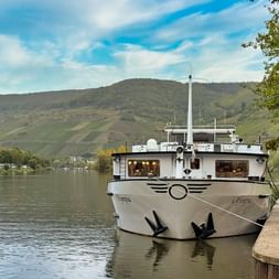 Weißes Flusskreuzfahrtschiff MS Olympia an einem Steinanleger festgemacht. Hügel mit Weinbergen und ein Dorf im Hintergrund unter blauem Himmel.