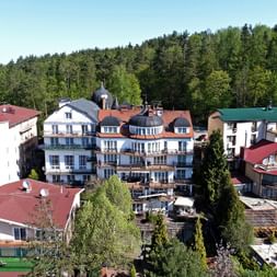 Luftaufnahme des Hotel Solar-Komplexes in Masuren mit weißen Gebäuden, roten Ziegeldächern und grünem Waldhintergrund. Außenterrasse im Vordergrund.