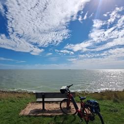 Rotes Tourenrad mit Taschen an einer Holzbank am grasbewachsenen Ufer des IJsselmeers unter blauem Himmel mit weißen Wolken.