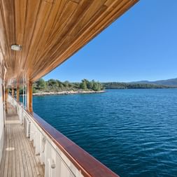 Blick vom Holzdeck der MY Magellan mit weißer Reling auf das blaue Adriatische Meer, grüne Küste und Berge unter klarem Himmel in Kroatien.
