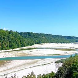 Breites Flussbett mit türkisfarbenen Wasserkanälen, die sich durch weiße Kiesbänke schlängeln, gesäumt von dichten grünen Wäldern unter blauem Himmel in Friaul.