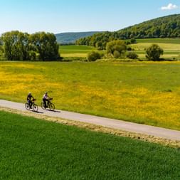 Zwei Radfahrer fahren auf einem asphaltierten Weg durch die hügelige Landschaft des Weserberglandes mit grünen Feldern, gelben Blumenwiesen und bewaldeten Hügeln.