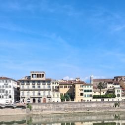 Reihe historischer italienischer Gebäude spiegelt sich im Arno in der Toskana, mit einem Kirchturm rechts unter blauem Himmel.