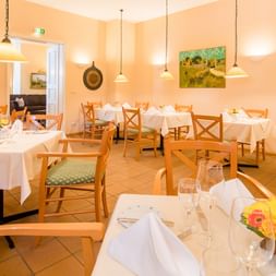 Helles Restaurant mit Holzstühlen, weißen Tischdecken, Pendelleuchten und bunten Gemälden an cremefarbenen Wänden im Hotel Geheimer Rat.