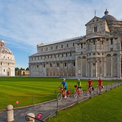 Radfahrer am Dom und Baptisterium von Pisa Radfahrergruppe auf Weg neben dem Dom von Pisa und rundem Baptisterium auf grüner Wiese. Historische weiße Marmorgebäude unter blauem Himmel.