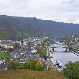 Luftaufnahme von Cochem mit Kirchturm, historischen Gebäuden und der Mosel mit Brücke und Booten. Bewaldete Hügel im Hintergrund unter bewölktem Himmel.