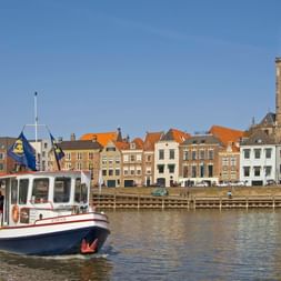 Weißes und blaues Fahrgastschiff auf Fluss mit historischer Deventer Skyline. Bunte Gebäude und hoher Kirchturm unter blauem Himmel.