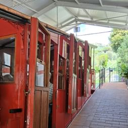 Rote hölzerne Standseilbahn mit offenen Fenstern an einer überdachten Bahnsteig in der Toskana, mit grünen Hügeln im Hintergrund.