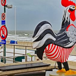 Große dekorative Hahnskulptur in Rot, Weiß und Schwarz an der Strandpromenade von De Haan. Blaue Strandkörbe und die Nordsee im Hintergrund.