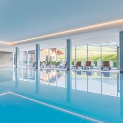 Modernes Hallenbad mit blauem Wasser, weißen Säulen und bodentiefen Fenstern mit Blick auf die Alpenlandschaft in Tirol.