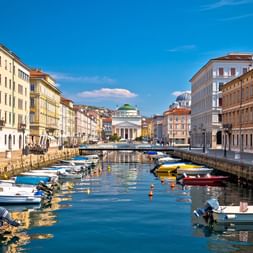 Canal Grande in Triest mit bunten Booten an Steinkai vertäut. Historische neoklassizistische Gebäude säumen beide Seiten unter blauem Himmel.