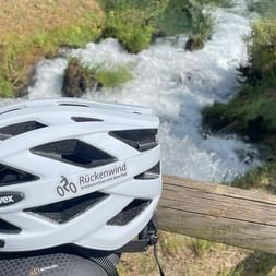 Weißer Uvex-Fahrradhelm mit Rückenwind-Logo auf Holzgeländer, mit rauschender Bergbach im Hintergrund.