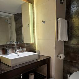Zeitgenössisches Hotelbadezimmer mit weißem Waschbecken, beleuchtetem Spiegel, dunklen Fliesen und weißen Handtüchern im Sheraton Essen.