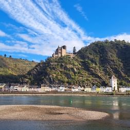 Burg Katz thront auf einem bewaldeten Hügel über einer Rheinstadt. Weiße Gebäude säumen das Flussufer mit einem Turm unter blauem Himmel.
