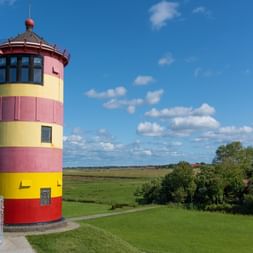 Pilsum Lighthouse in East Frisia with distinctive red, yellow and pink horizontal stripes, surrounded by green meadows under blue sky.