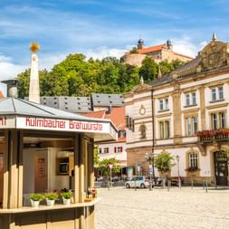 Kopfsteinpflasterplatz in Kulmbach mit Kulmbacher Brauverein Bierstand und historischen Barockgebäuden. Burg auf Hügel im Hintergrund.