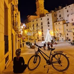 Radfahrerin mit Helm sitzt neben ihrem Fahrrad auf einem gepflasterten Platz in Valencia bei Nacht und betrachtet beleuchtete Gebäude und Weihnachtsdekoration.