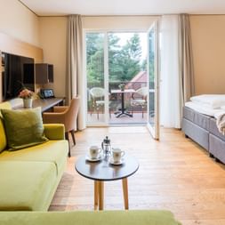 Modernes Hotelzimmer mit grünem Sofa, Doppelbett, TV und Balkontür mit Blick auf Sitzbereich und Garten.