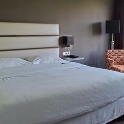 Modernes Hotelzimmer mit Doppelbett, weißer Bettwäsche, gepolstertem Kopfteil, gemustertem Sessel und großem Fenster mit Blick ins Grüne.