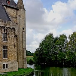 Steinernes mittelalterliches Schloss in Brügge mit rundem Turm und vielen Fenstern, umgeben von Wasser und grünen Bäumen unter bewölktem Himmel.