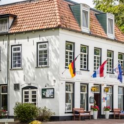 Weißes zweistöckiges Hotelgebäude mit rotem Ziegeldach und Dachgauben. Mehrere Flaggen, darunter deutsche und niederländische, hängen an der Fassade.
