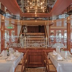 Elegantes Restaurant an Bord der MS Switzerland mit weiß gedeckten Tischen, Holzstühlen und einem zentralen Klavier. Warme Beleuchtung und Fenster.
