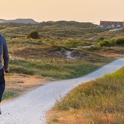 Radfahrer mit rotem Rucksack schiebt Fahrrad auf gewundenem Weg durch grasbewachsene Dünen auf Terschelling bei Sonnenuntergang.