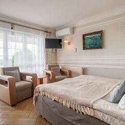 Elegantes Hotelzimmer in Masuren mit Doppelbett, zwei braunen Sesseln, Balkontüren, Wandfernseher und klassischem weißen Interieur.
