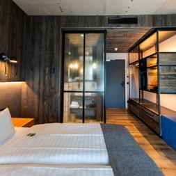 Zeitgenössisches Hotelzimmer mit Doppelbett, dunklen Holzwänden, Glas-Badabtrennung und blauem Sofa im Hotel Zollhaus in Rastede.
